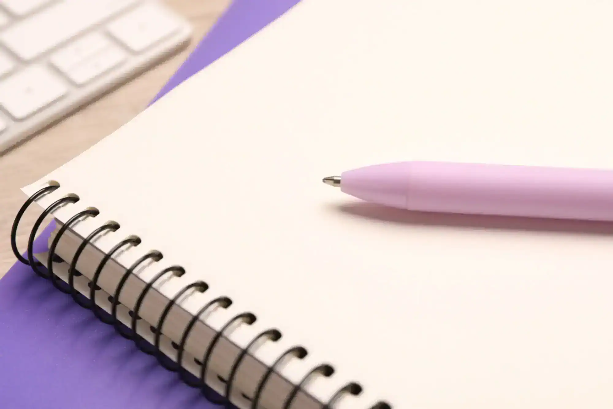 Pink pen resting on a blank notebook page with a purple cover, with a keyboard in the background.
