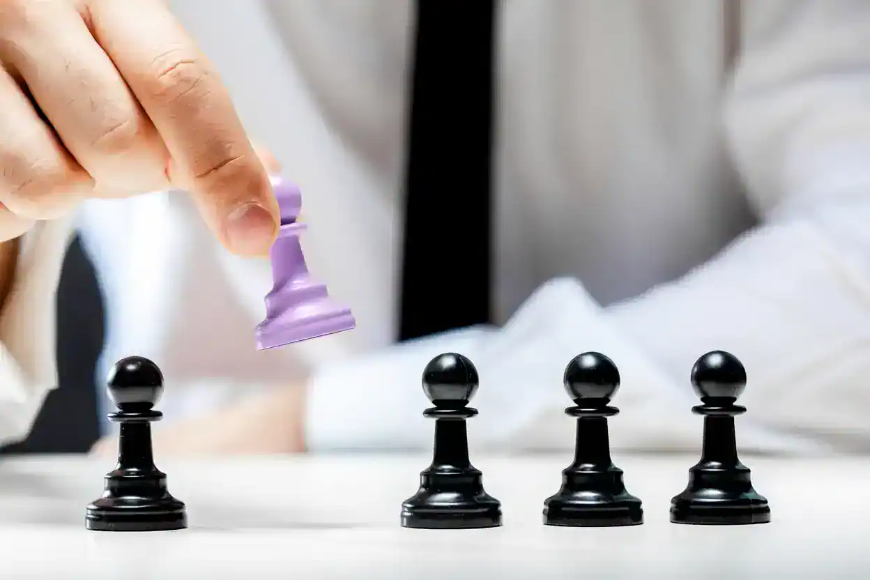 Hand moving a lavender chess piece above a row of black pawns on a table.