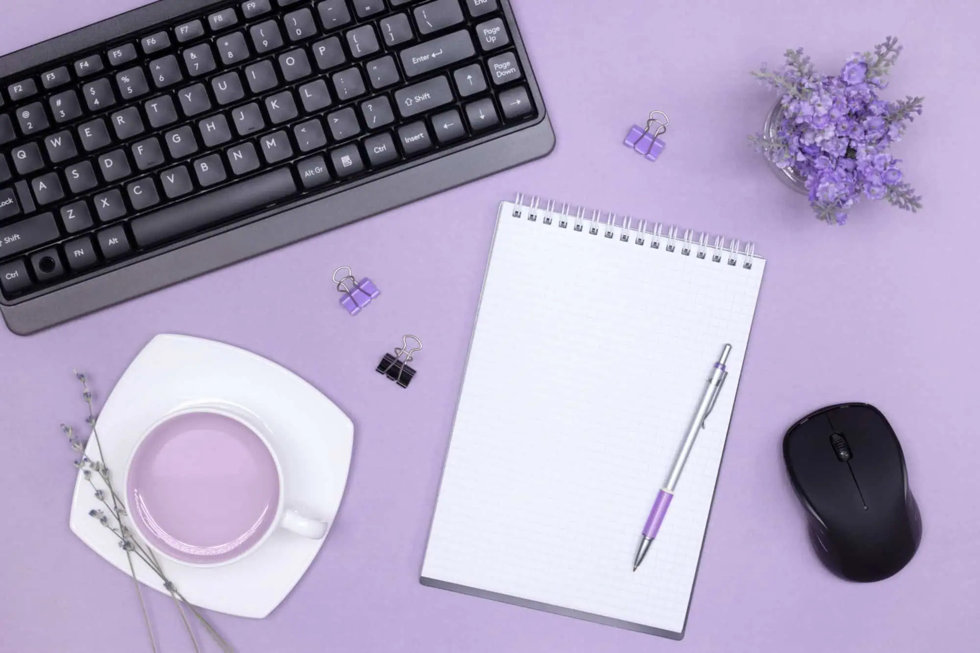 Flat lay of a lavender desk with a keyboard, blank notepad with pen, mouse, and a cup of purple tea.