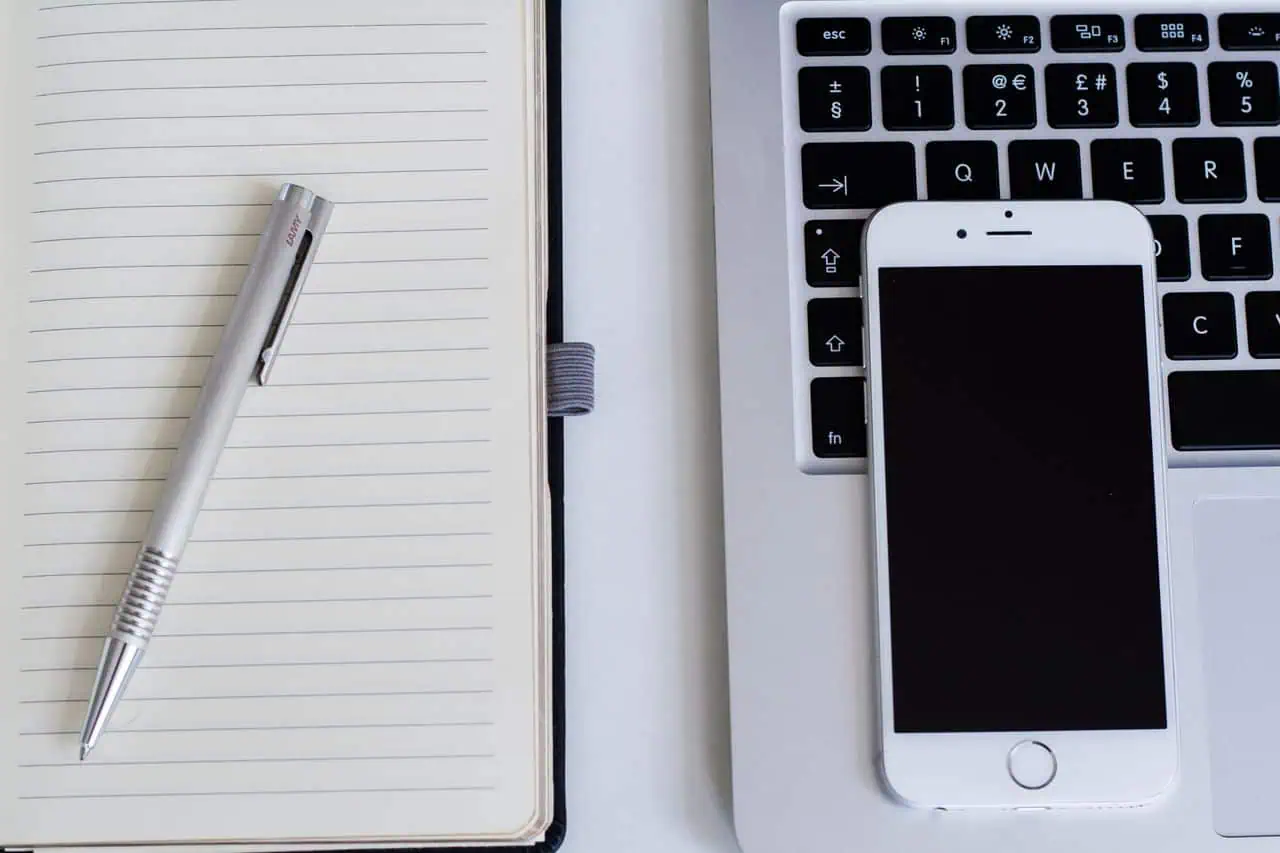 Close-up of a notebook, pen, smartphone, and laptop on a modern workspace.